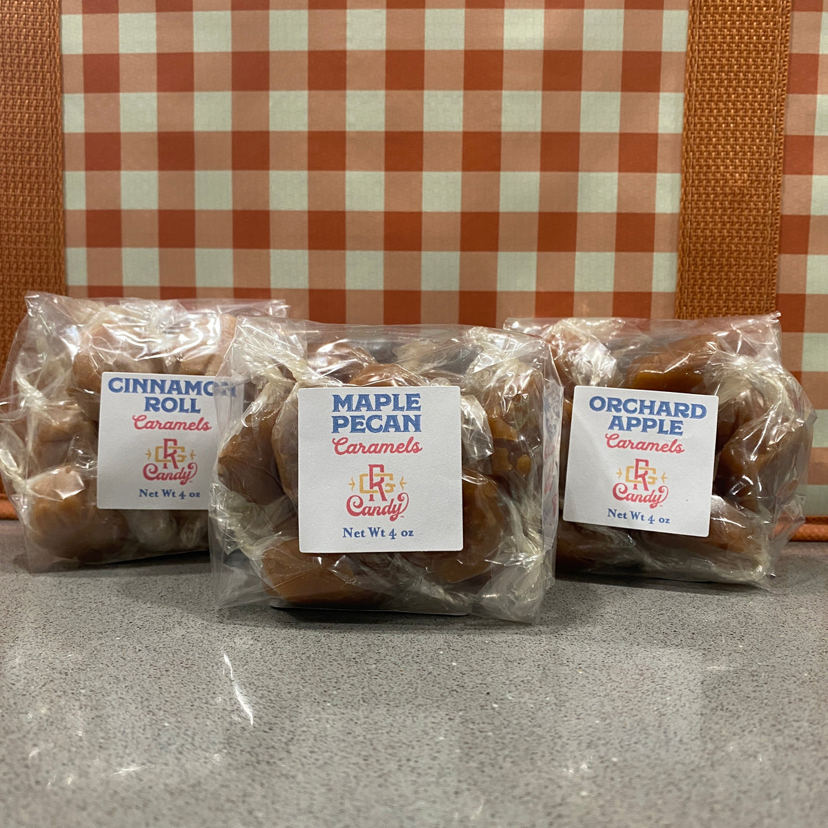 Three individually wrapped packages of B&B Candy gourmet caramels lined up on a gray countertop. The packages are labeled Cinnamon Roll Roll, Maple Pecan, and Orchard Apple, set against a fall colored checkered background.