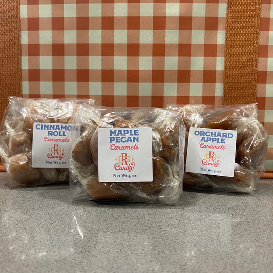 Three individually wrapped packages of B&B Candy gourmet caramels lined up on a gray countertop. The packages are labeled Cinnamon Roll Roll, Maple Pecan, and Orchard Apple, set against a fall colored checkered background.